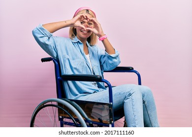 Beautiful blonde woman sitting on wheelchair doing heart shape with hand and fingers smiling looking through sign  - Powered by Shutterstock