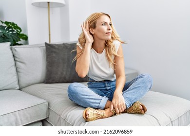 Beautiful Blonde Woman Sitting On The Sofa At Home Smiling With Hand Over Ear Listening An Hearing To Rumor Or Gossip. Deafness Concept. 