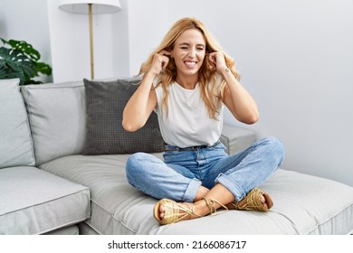 Beautiful blonde woman sitting on the sofa at home covering ears with fingers with annoyed expression for the noise of loud music. deaf concept. 