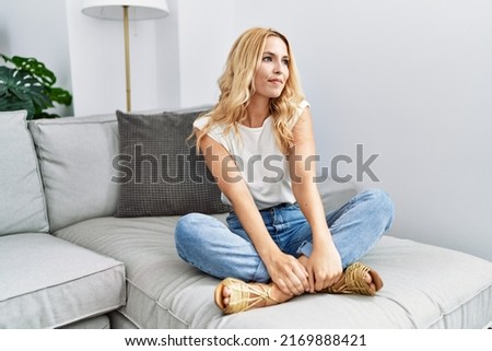 Beautiful blonde woman sitting on the sofa at home looking away to side with smile on face, natural expression. laughing confident. 