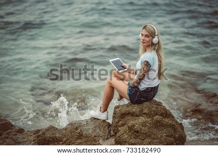Beautiful blonde woman sitting on a rock by the sea and listening to music on stylish headphones on the tablet. hands with a tattoo. The style of summer holiday. Modern girl