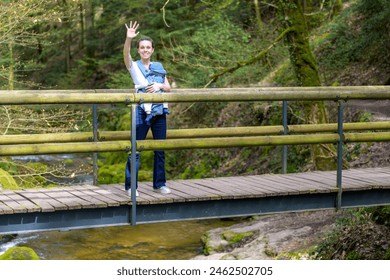 Beautiful blonde woman standing on a bridge in nature with her little baby and waving to the camera - Powered by Shutterstock