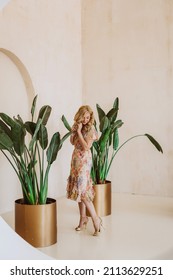 A beautiful blonde woman in a summer dress with a floral print against the background of large houseplants Strelitzia in golden flowerpots.
