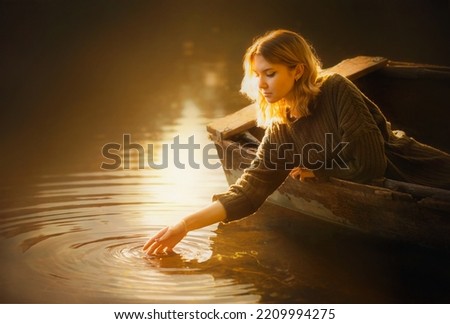  A beautiful blonde woman in a sweater sits in an old fishing boat and touches the surface of the river water with her hand, illuminated by the light of the rising sun. Tranquility and a fairy tale.