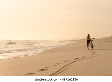 Beautiful blonde woman walking with her bicycle down a quiet beach at sunset.