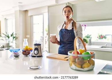 Beautiful Blonde Woman Wearing Apron Drinking Smoothie At The Kitchen Scared And Amazed With Open Mouth For Surprise, Disbelief Face 