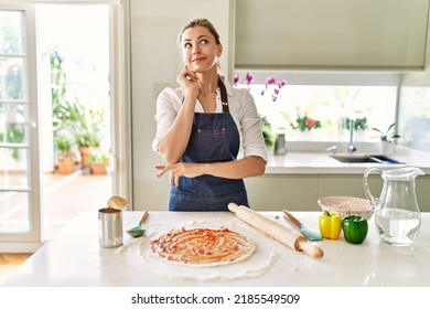 Beautiful blonde woman wearing apron cooking pizza serious face thinking about question with hand on chin, thoughtful about confusing idea 