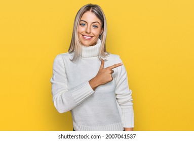 Beautiful blonde woman wearing casual turtleneck sweater cheerful with a smile of face pointing with hand and finger up to the side with happy and natural expression on face 