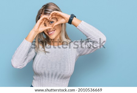 Beautiful blonde woman wearing casual clothes doing heart shape with hand and fingers smiling looking through sign 