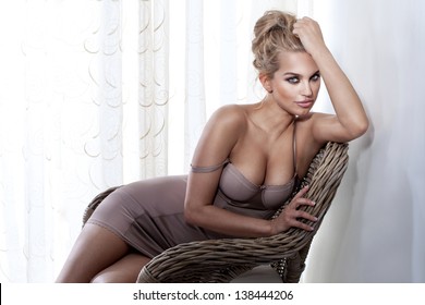 Beautiful blonde woman wearing fashionable lingerie, sitting on wicker chair in bright room.