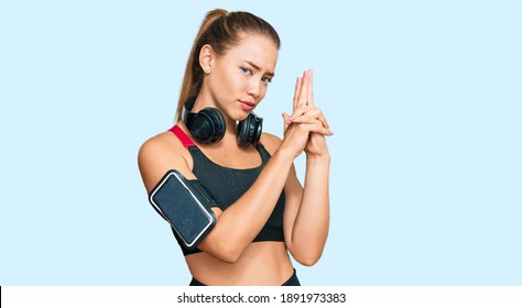 Beautiful blonde woman wearing gym clothes and using headphones holding symbolic gun with hand gesture, playing killing shooting weapons, angry face 