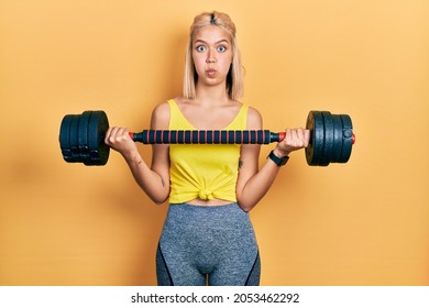 Beautiful blonde woman wearing sportswear using dumbbells puffing cheeks with funny face. mouth inflated with air, catching air. 