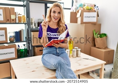 Beautiful blonde woman wearing volunteer t shirt at donations stand taking notes surprised with an idea or question pointing finger with happy face, number one 