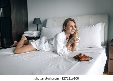 Beautiful blonde woman in white coat with white smile having breakfast in a hotel room. Woman is having breakfast with croissant in the morning!