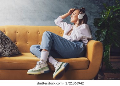 Schöne Blondine in weißen Kopfhörern sitzt auf einem gelben Sofa und hört gerne Musik, Podcast.