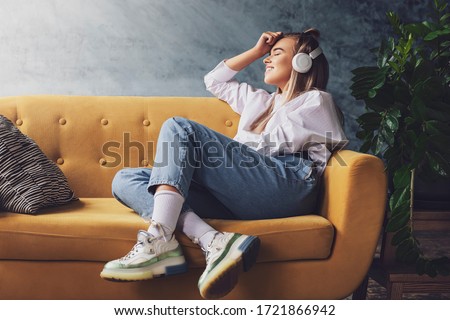 Beautiful blonde woman in white headphones sits on a yellow sofa and enjoys listening to music, podcast.