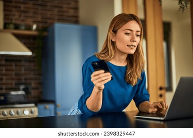 A beautiful blonde woman working from home, using a laptop and a mobile phone. - Powered by Shutterstock