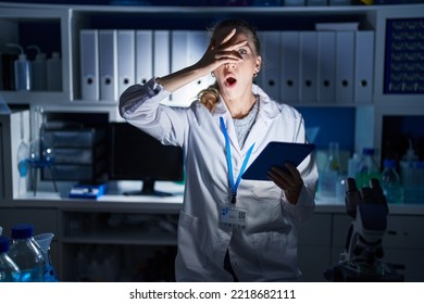 Beautiful Blonde Woman Working At Scientist Laboratory Late At Night Peeking In Shock Covering Face And Eyes With Hand, Looking Through Fingers With Embarrassed Expression. 