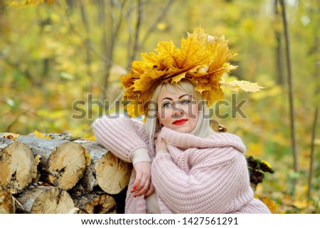 Beautiful blonde in a wreath of autumn leaves, sitting in a gentle jumper in the woods leaning on a log, thoughtfully and smiling. Woman's size plus xxl