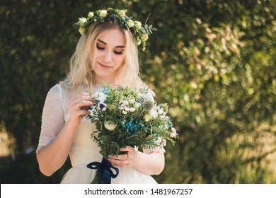 Beautiful Blonde Young Bride Portrait With Flower Bouquet And Wreath On Her Head In White Wedding Dress Outdoor In Summer