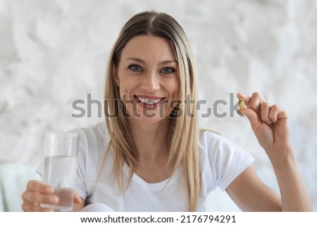Beautiful blonde young lady holding pill and glass of water, smiling at camera, closeup photo of happy attractive lady taking supplement vitamin D after waking up in the morning, copy space