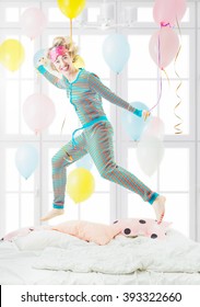 Beautiful blonde young woman having fun jumping on the bed in striped cool pijama sleepwear. Crazy happy morning mood. Good morning world! Sunny bedrom with baloons