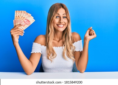 Beautiful blonde young woman holding 100 mexican pesos banknotes screaming proud, celebrating victory and success very excited with raised arm 