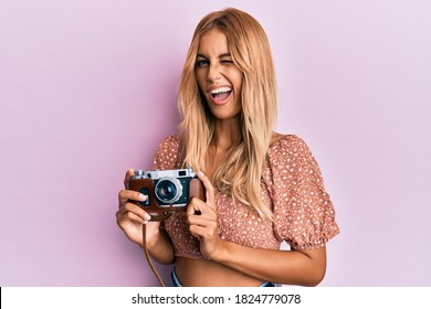 Beautiful blonde young woman holding vintage camera winking looking at the camera with sexy expression, cheerful and happy face. 