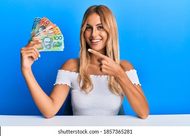 Beautiful blonde young woman holding australian dollars smiling happy pointing with hand and finger 