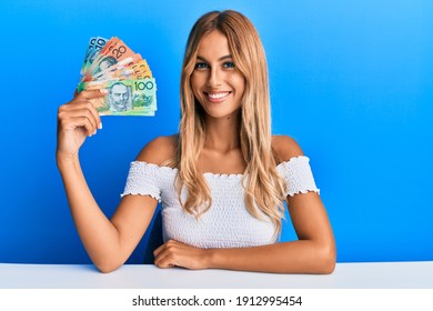 Beautiful blonde young woman holding australian dollars looking positive and happy standing and smiling with a confident smile showing teeth 