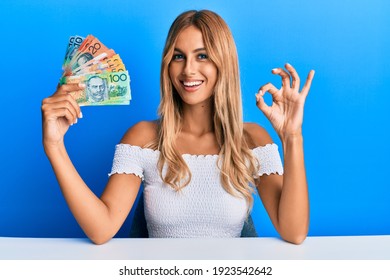 Beautiful blonde young woman holding australian dollars doing ok sign with fingers, smiling friendly gesturing excellent symbol 