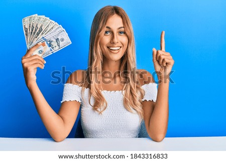 Beautiful blonde young woman holding dollars smiling with an idea or question pointing finger with happy face, number one 