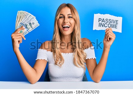 Beautiful blonde young woman holding dollars and passive income text smiling and laughing hard out loud because funny crazy joke. 