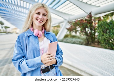 Beautiful blonde young woman smiling happy holding book and wearing headphones outdoors on a sunny day - Shutterstock ID 2139327643