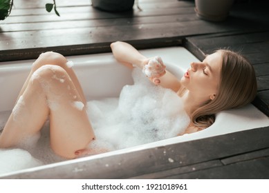 Beautiful blonde young woman taking bath with bubbles in bathroom interior with plenty green plans. Lifestyle wellness concept. Treating yourself and wellbeing