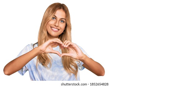 Beautiful Blonde Young Woman Wearing Casual Tie Dye Shirt Smiling In Love Showing Heart Symbol And Shape With Hands. Romantic Concept. 