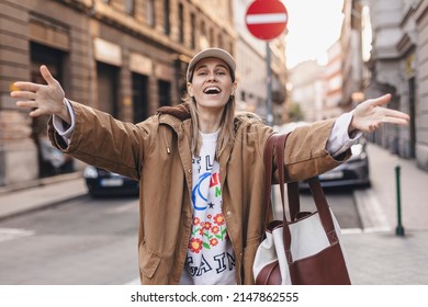 Beautiful blonde young woman wearing brown trench coat and white cap looking at the camera smiling with open arms for hug, cheerful expression embracing happiness, happy meeting of friends or family.