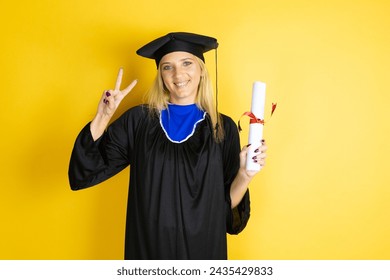 Beautiful blonde young woman wearing graduation cap and ceremony robe showing and pointing up with fingers number two while smiling confident and happy