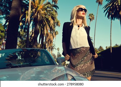 Beautiful blonde young woman wearing fashionable clothes, black cardigan posing next to a car on the street with palm trees. Fashion photo