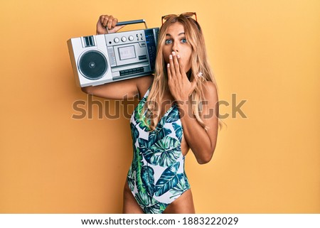 Beautiful blonde young woman wearing swimsuit and holding boombox covering mouth with hand, shocked and afraid for mistake. surprised expression 