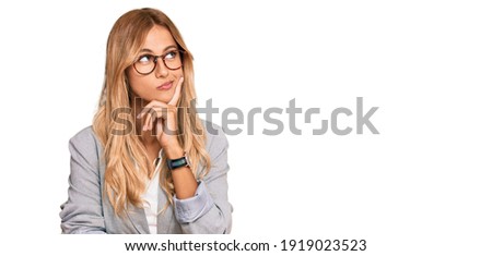 Beautiful blonde young woman wearing business clothes with hand on chin thinking about question, pensive expression. smiling with thoughtful face. doubt concept. 