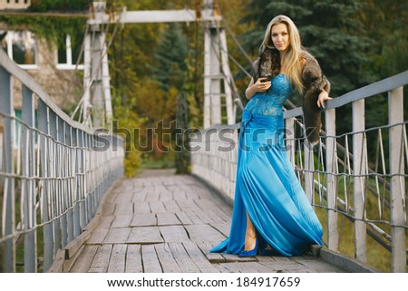 Beautiful blondy young woman wearing elegant blue dress standing on a bridge in the park 