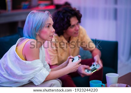 Beautiful blue haired girl and young guy holding gaming contorllers playing video games at home.