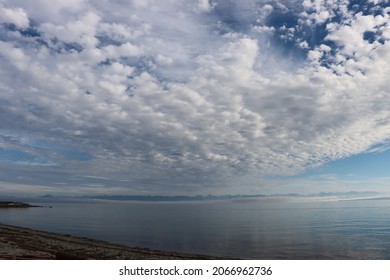 A Beautiful Blue Sky With Copious Clouds Over Calm Ocean Waters. Large, White, Fluffy Clouds Disappear Into The Horizon. Layers Of Cloud Shapes Blanket The Sky And Are Reflected Into Still Waters.