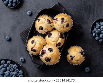 Beautiful blueberry muffins in black baking form with fresh blueberries on dark background. Delicious fruit cakes, dessert or bakery. Rustic style. Culinary magazine cover. Postcard design. Top view.