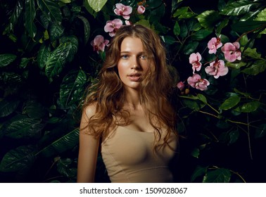 A beautiful blue-eyed long-haired brown-haired woman stands on a background of exotic plants and pink orchids.Beauty, fashion, cosmetology, natural, nature, natural, spa, detox, plants.