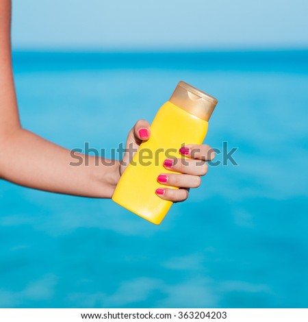 Beautiful body young woman in pink heart patterned yellow bikini and pink nails holding yellow bottled sunscreen with azure blue ocean sea beach background