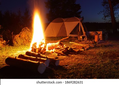 Beautiful bonfire with bright orange flame light, firewood and camp tent on camp site at calm summer night, romantic lifestyle outdoor tourism vacation on nature landscape in Russia on empty glade