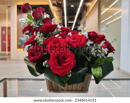 Beautiful bouquet of red roses, white daisies and white chrysanthemums in a bamboo basket put on a long glass table with office interior on blurred background. Used for room decoration