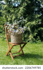 Beautiful Bouquet Of Wildflowers In Wicker Basket On Wooden Garden Seat. Comfort Lounge Chair With Decoration On Green Lawn In Backyard At Summer Day